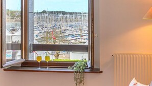A room with a view of a marina, a window sill with two glasses of yellow drink, a hanging plant, and a cushion with text. - Le Quai by Interhome (La Trinité Sur Mer)