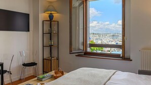 A bedroom with a bed, a nightstand, a lamp, a shelf, and a window with a view of a marina. - Le Quai by Interhome (La Trinité Sur Mer)