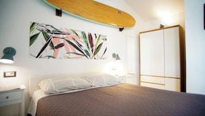 A bedroom with a bed, a wall-mounted surfboard, a wardrobe, and a wall with a tropical leaf pattern. - Sa Domu de Jana - Studio apartment with garden and panoramic view of Poetto (Quartu Sant'Elena)