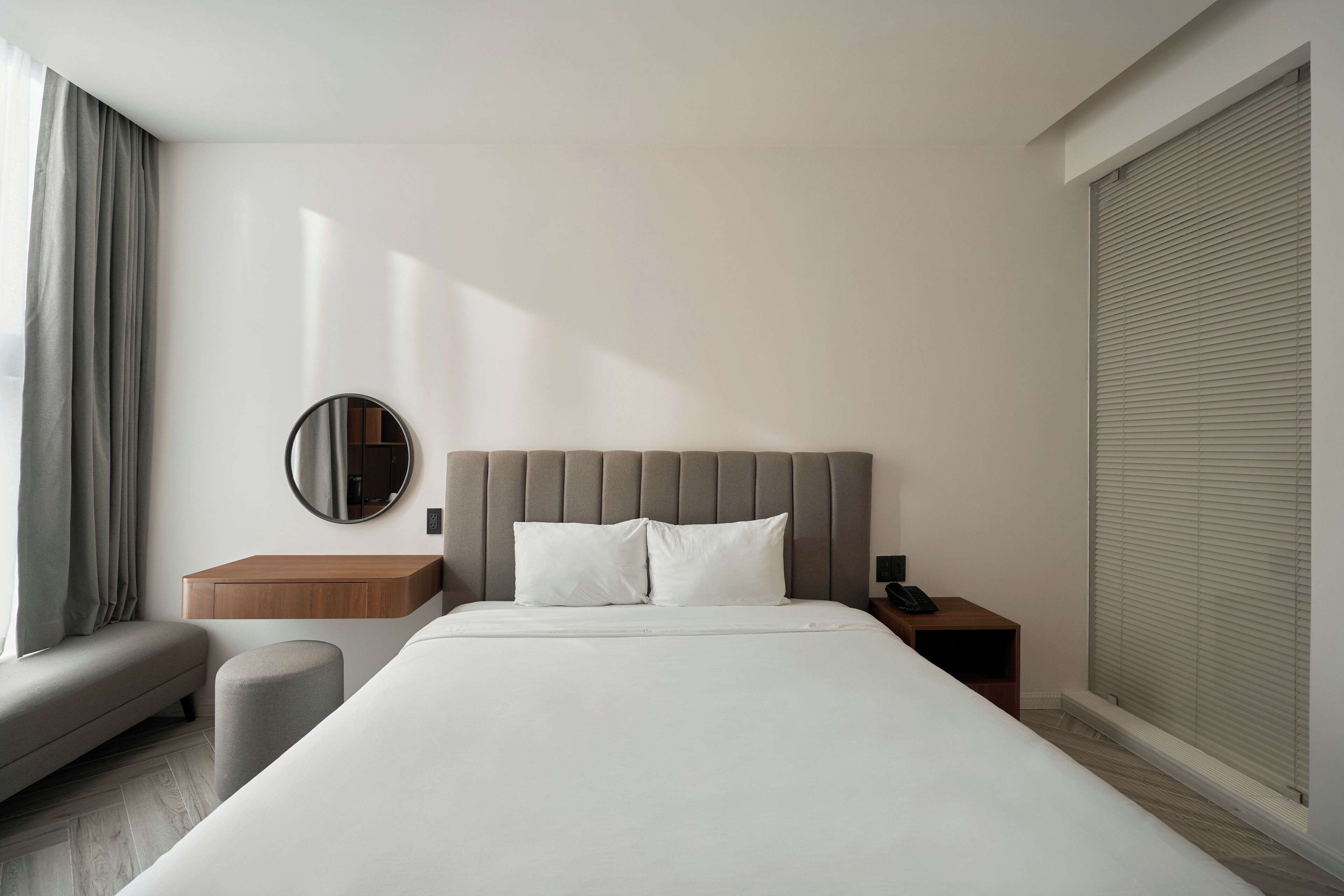 Une chambre d’hôtel moderne avec un grand lit, une table de chevet, un miroir rond et une fenêtre avec des rideaux.