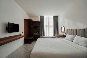 Una habitación de hotel moderna con una cama grande, un televisor montado en la pared, un armario de madera y una ventana con cortinas.