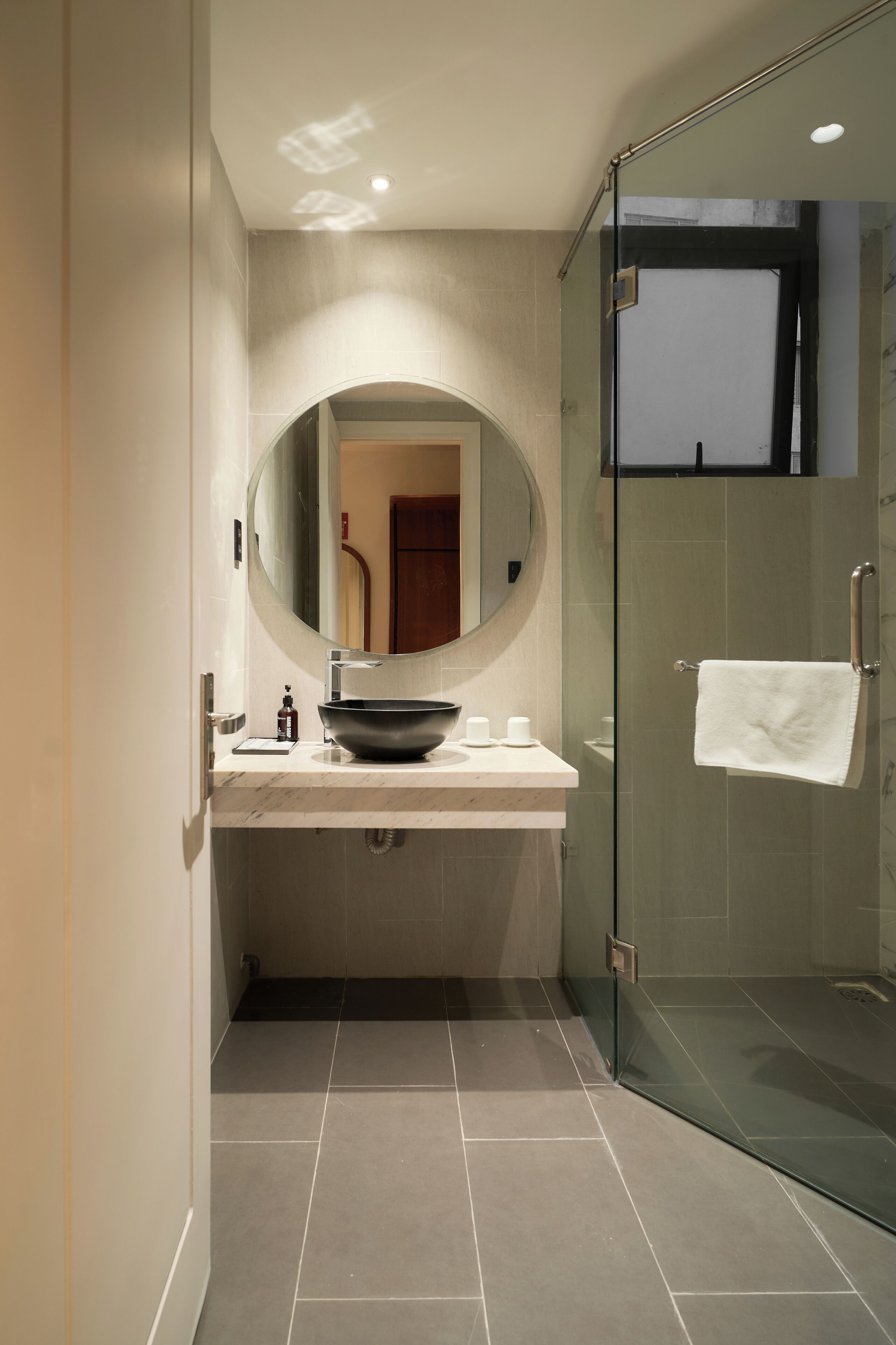A modern bathroom with a round mirror, a vessel sink, a glass-enclosed shower, and a towel hanging on the door.