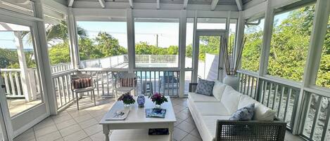 A covered outdoor area with a white ceiling, white walls, and a white floor. There is a white sofa, a wicker chair, and a white table with decorative items.