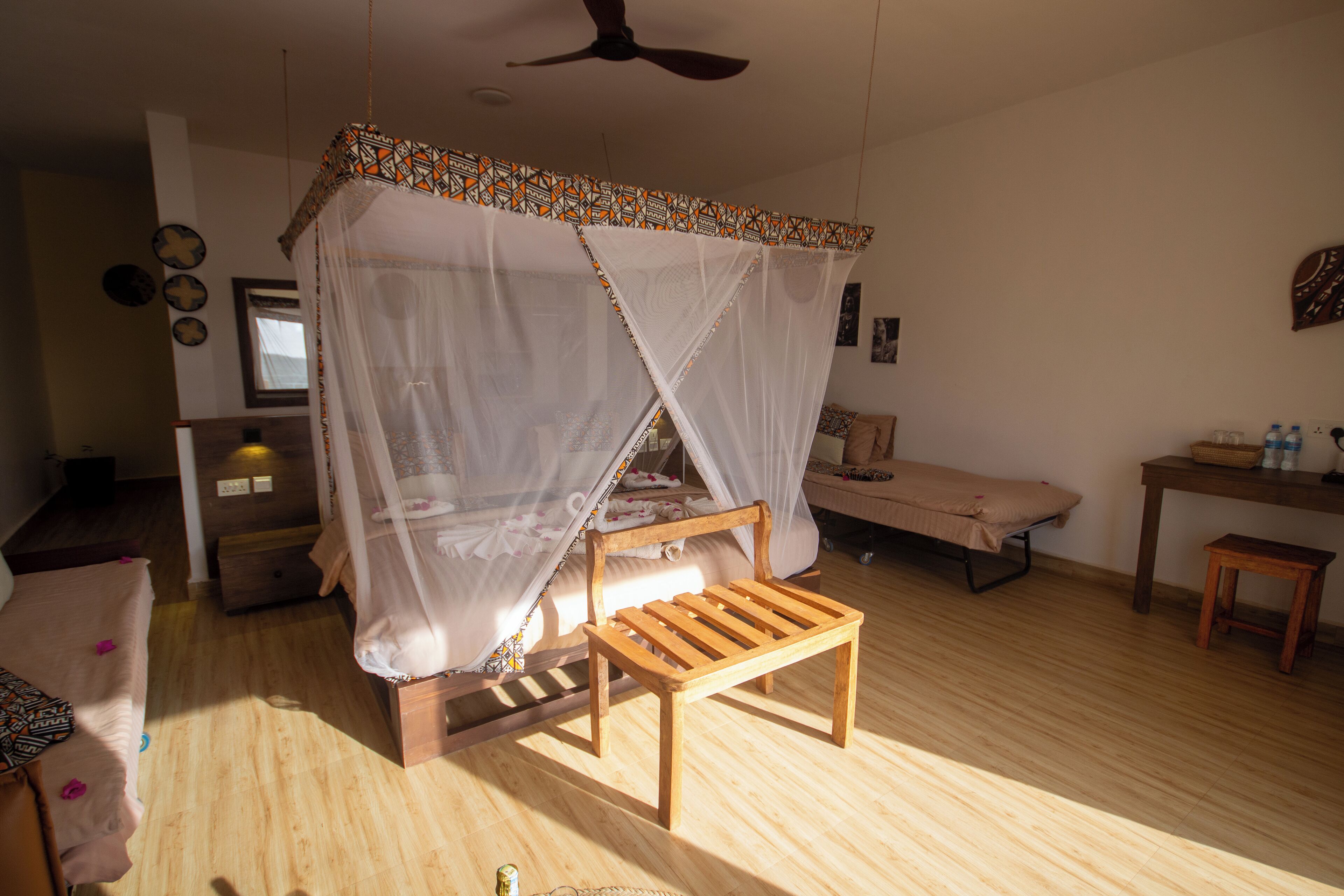 A room with a wooden bed under a canopy, a small wooden bench, a desk with a chair, and a ceiling fan.