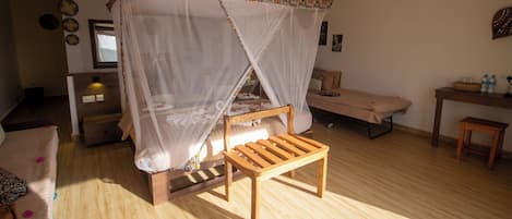 A room with a wooden bed under a canopy, a small wooden bench, a desk with a chair, and a ceiling fan.