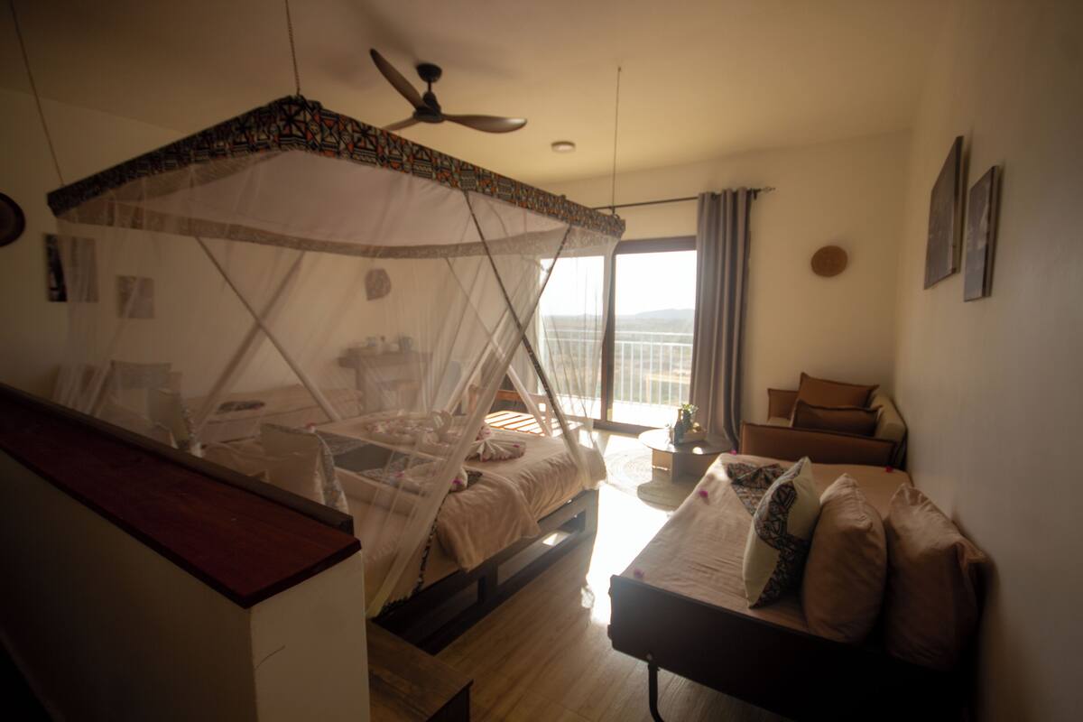 Ein Schlafzimmer mit einem Bett unter einem Moskitonetz, einem Deckenventilator, einer Couch und Blick auf die Stadt.