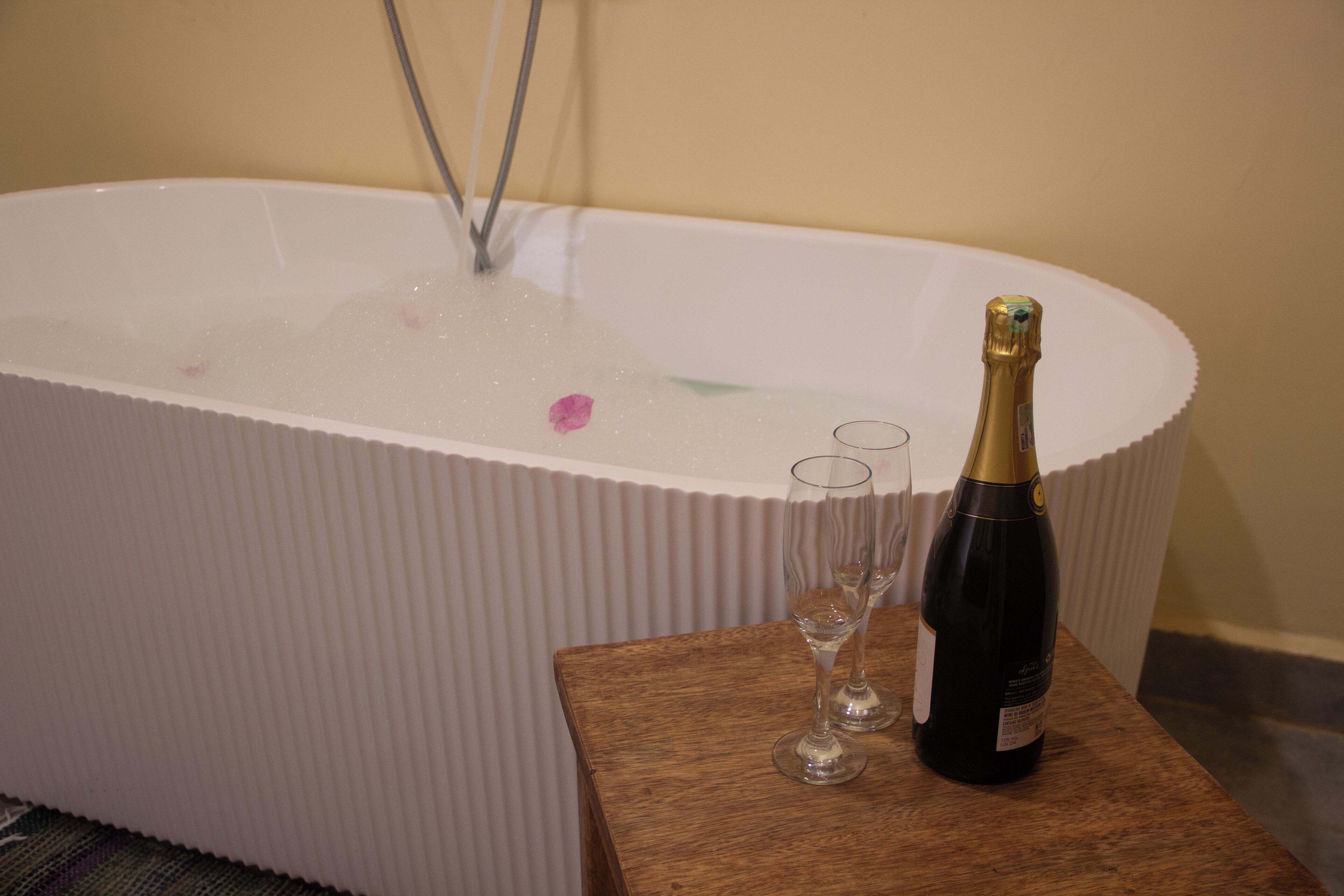 A bathtub with bubbles, a bottle of champagne, and two glasses on a wooden table.
