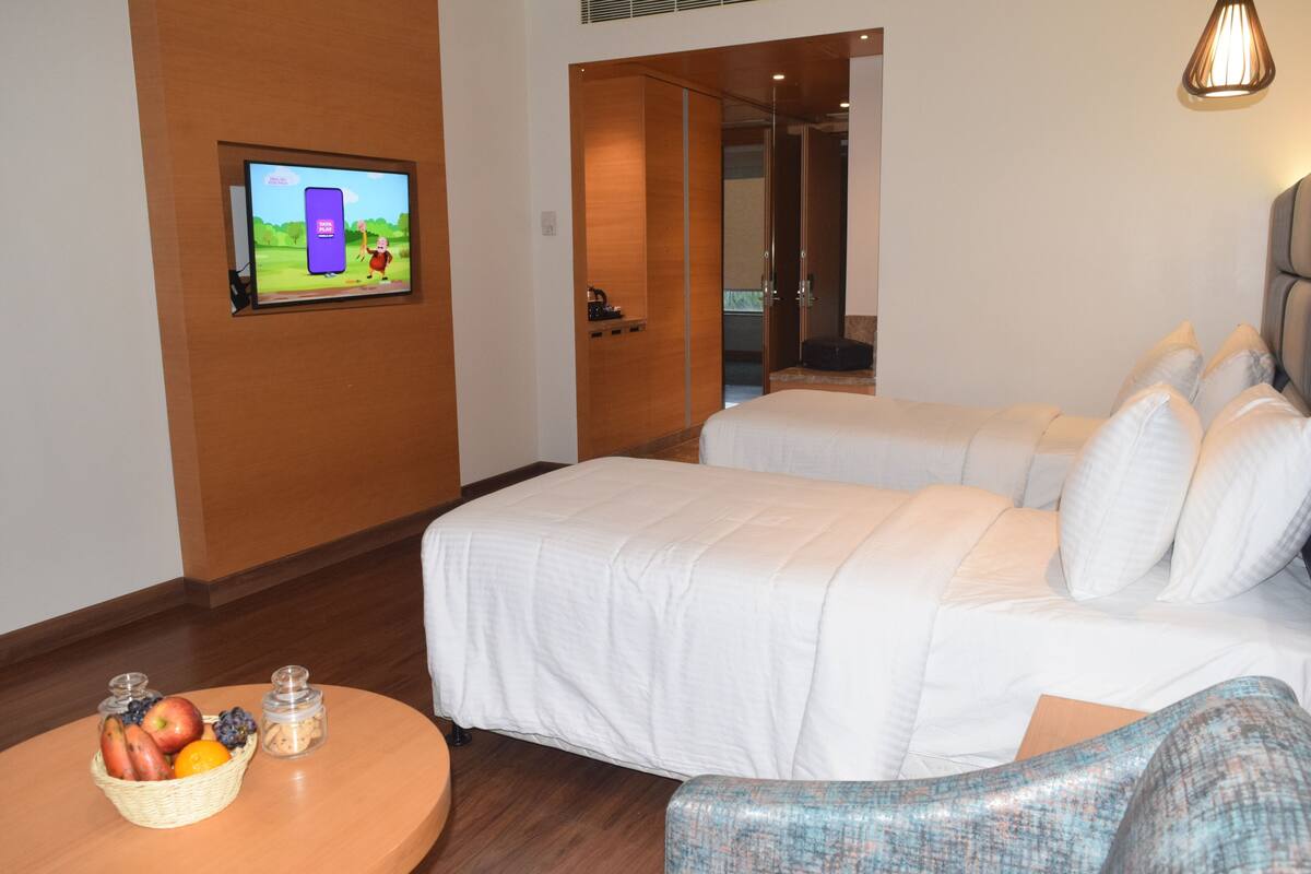 Une chambre d’hôtel avec deux lits, un téléviseur fixé au mur, une petite table avec des fruits, et une porte donnant sur une salle de bain.