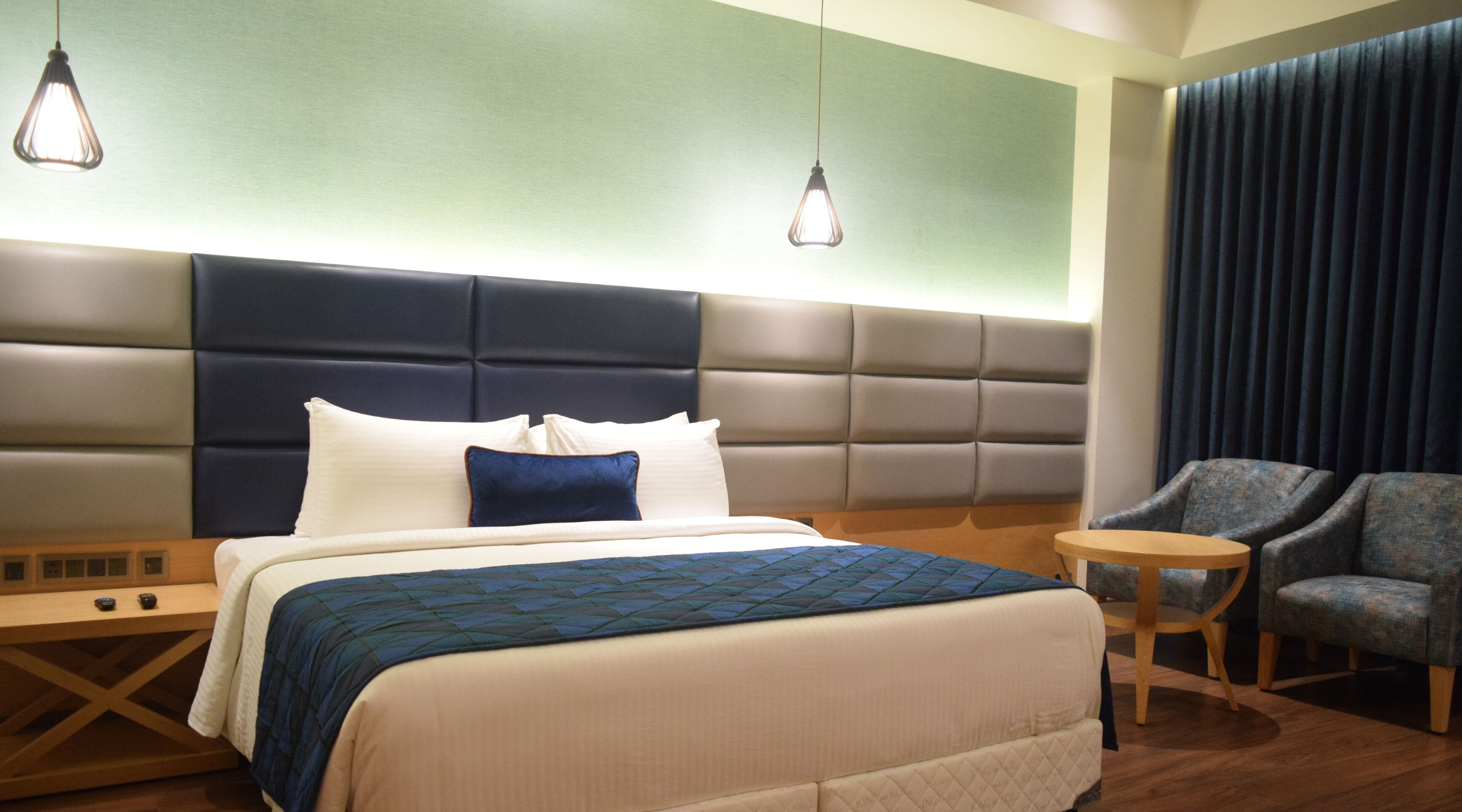 A hotel room with a large bed, two bedside tables, a seating area with a chair, and a headboard with built-in lighting.