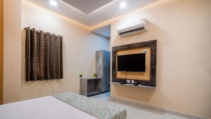 A hotel room with a bed, a TV mounted on the wall, an air conditioning unit, and a small seating area with a plant. - Hotel Chotiwala – 10 Mins from Kashi Vishwanath Temple (Varanasi)