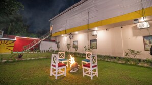 A building with a yellow and white exterior, a fire pit in the center, and two white chairs with red cushions. - Hotel Chotiwala – 10 Mins from Kashi Vishwanath Temple (Varanasi)