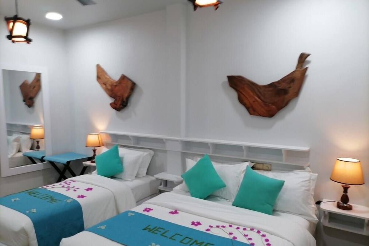 A hotel room with two beds, each with a 'WELCOME' sign, teal pillows, and a blue bedspread. There are wall-mounted wooden art pieces and a mirror.