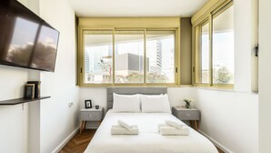 Un dormitori modern amb un llit gran, un televisor de pantalla plana fixat a la paret i una finestra amb vista a edificis.