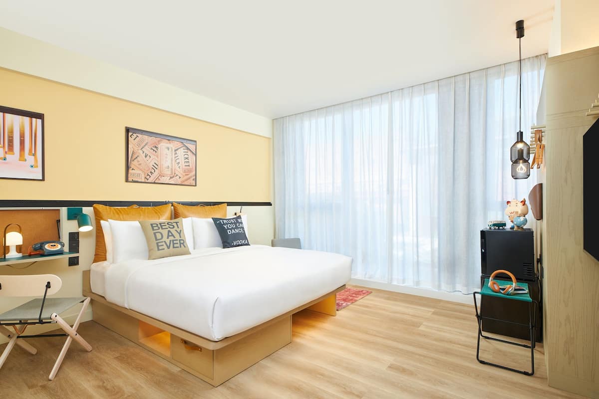 A modern hotel room with a large bed, a desk with a chair, a small table with a lamp, and a large window with curtains.