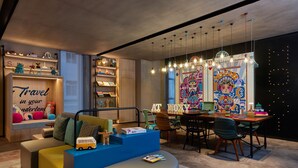 A modern living room with a sofa, ottoman, and a dining area with chairs and a table. There are shelves with decorative items and a wall with colorful artwork. - Moxy Xi'an Downtown (Xi'an)