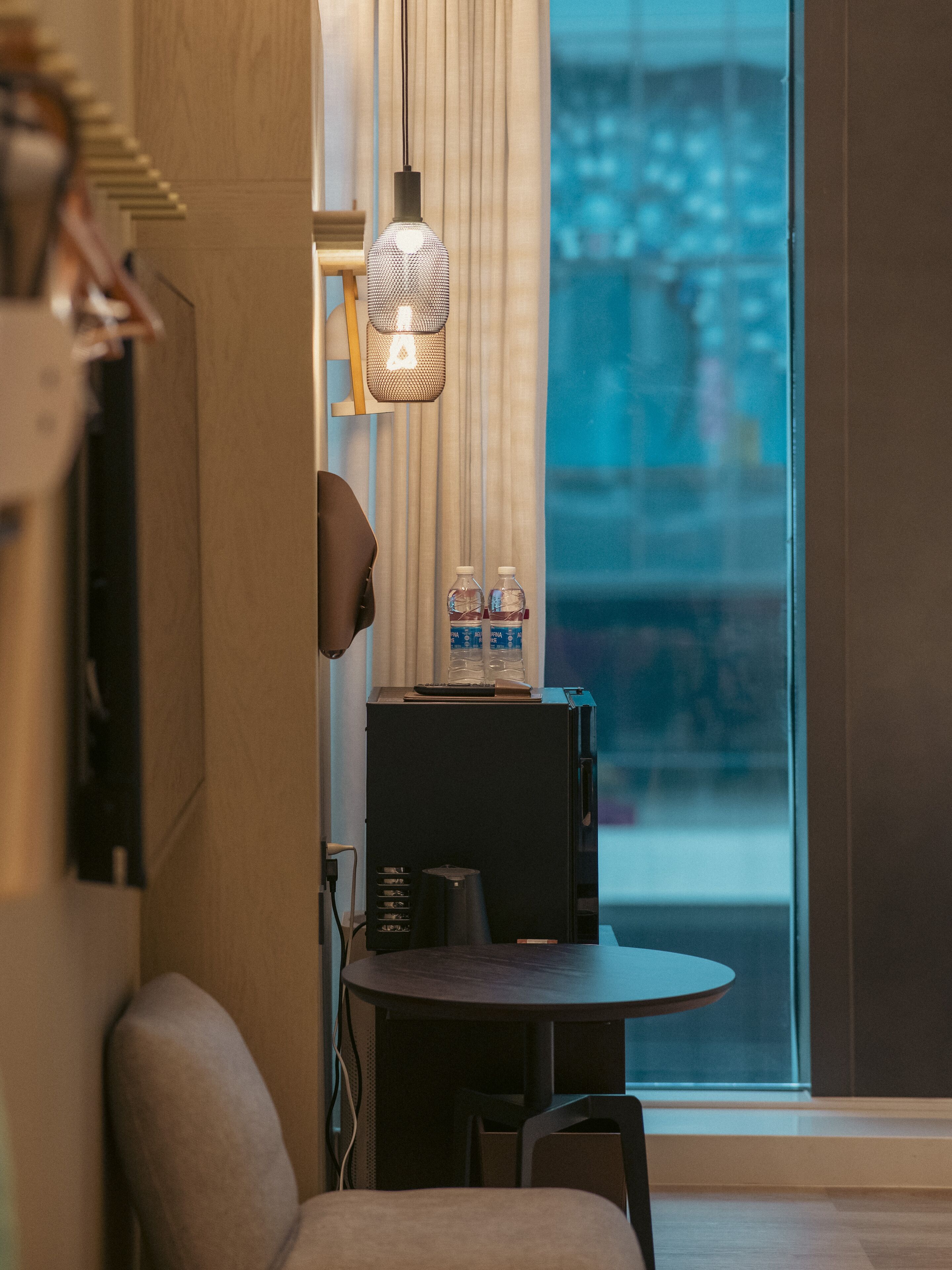 Habitación de hotel moderna con comedor que presenta una mesa redonda, dos sillas, una cafetera negra y un dispensador de agua. Una lámpara colgante ilumina la mesa, y un amplio ventanal ofrece vistas de la ciudad.