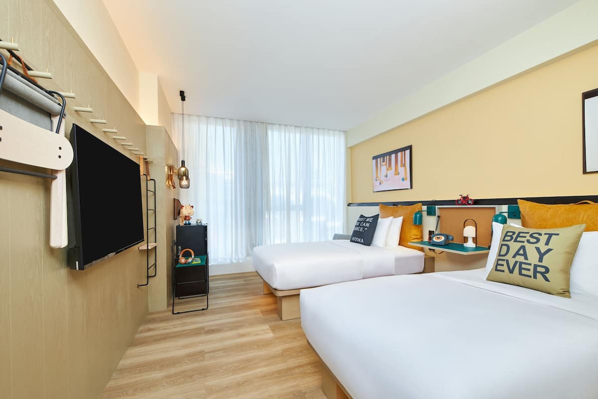 A hotel room with a large flat-screen TV, two beds, a desk with a chair, a small table with a lamp, and a window with sheer curtains.