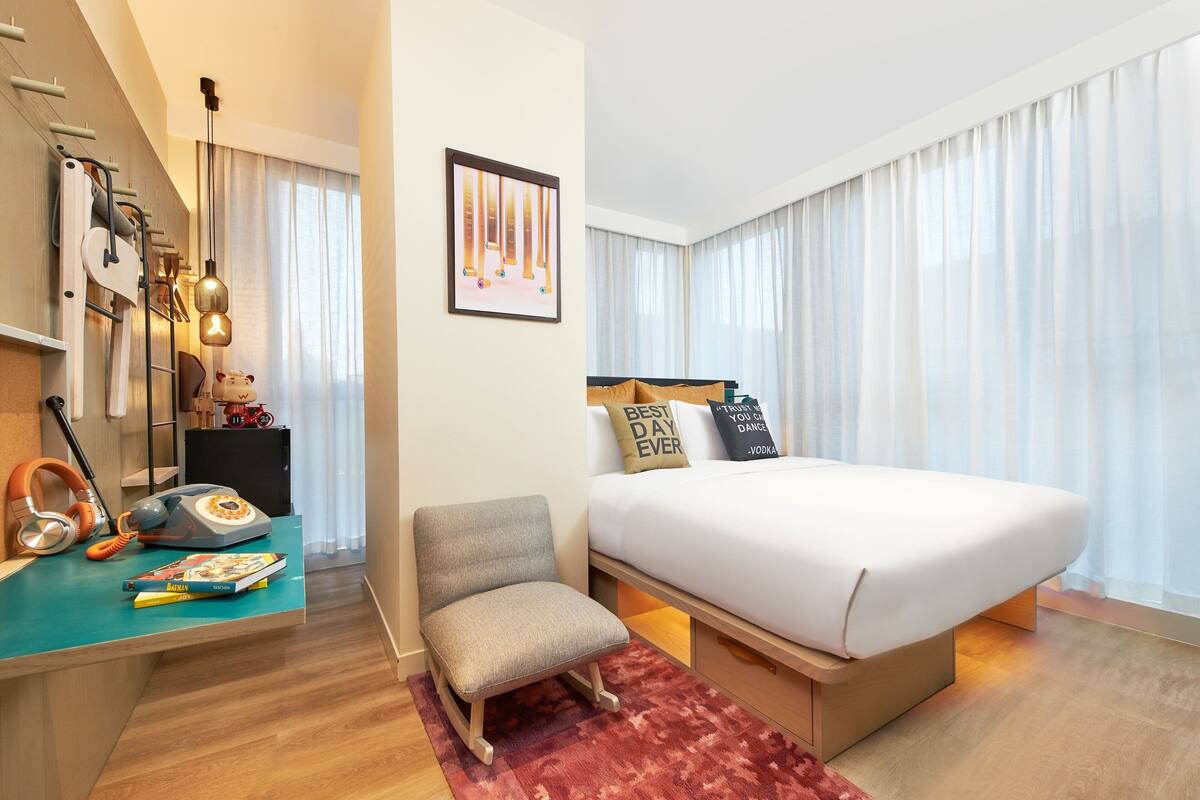 A modern hotel room with a large bed, a desk with a telephone and books, a chair, and a large window with curtains.