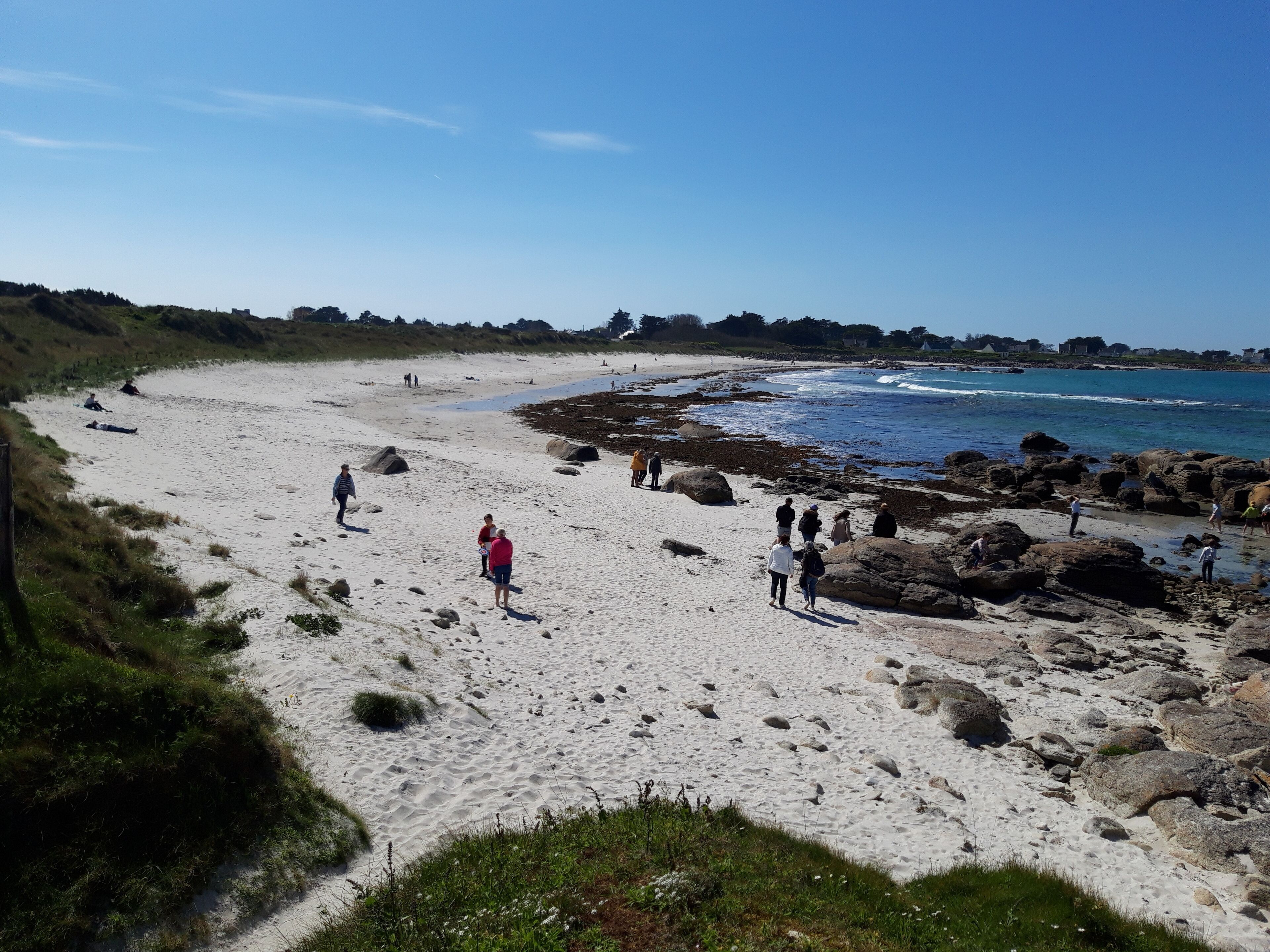 A sandy beach with people walking and standing, rocky areas near the water, and greenery in the background.