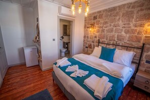 A bedroom with a stone wall, a bed with white linens and teal bedding, a small bedside table, and a wooden floor. - Cunda Alesta Otel (Ayvalik)