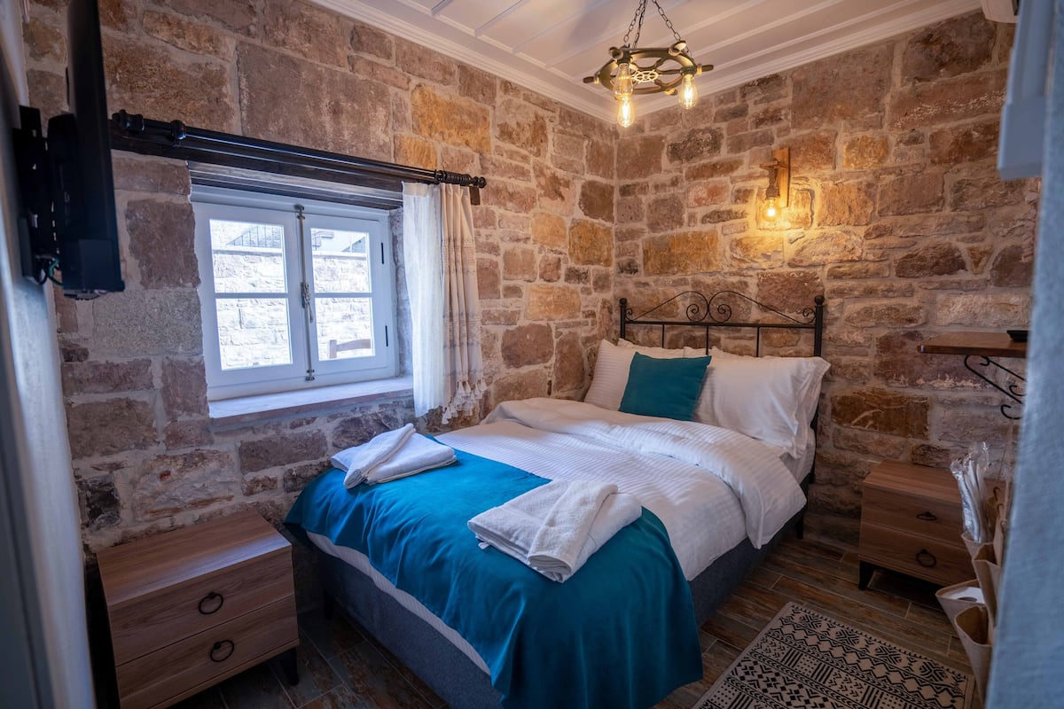 Quarto com parede de pedra, cama com roupa de cama branca e verde-água, mesa de cabeceira de madeira e luminária pendente.