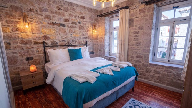 A bedroom with a stone wall, a bed with white and teal bedding, a wooden floor, and a window with curtains. - Cunda Alesta Otel (Ayvalik)