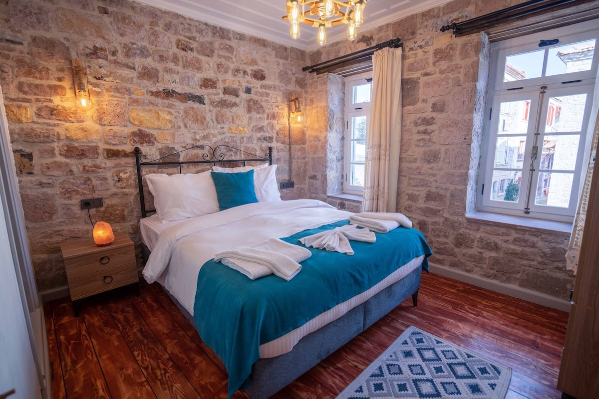 Quarto com parede de pedra, cama com roupa de cama branca e azul turquesa, piso de madeira e janela com cortinas.