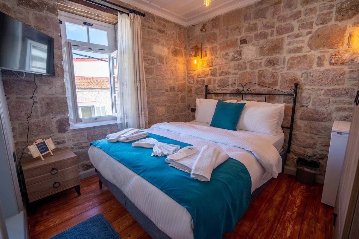 Quarto com parede de pedra, cama com roupa de cama branca e azul-turquesa, mesa de cabeceira de madeira com telefone e uma pequena placa, e janela com cortinas.
