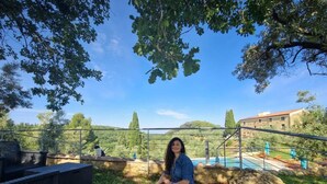 Une personne est assise sur un banc dans un jardin, avec une piscine et un bâtiment en arrière-plan.