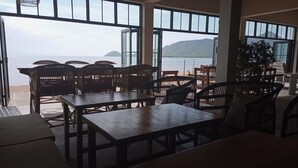 Un restaurant situé en bord de mer, doté de grandes fenêtres, de meubles en osier et offrant une vue sur la mer et les montagnes.