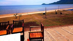 Un espace aménagé avec des chaises et une table en bois, situé au bord de la plage, d’où l’on peut admirer une mer calme et des collines lointaines.