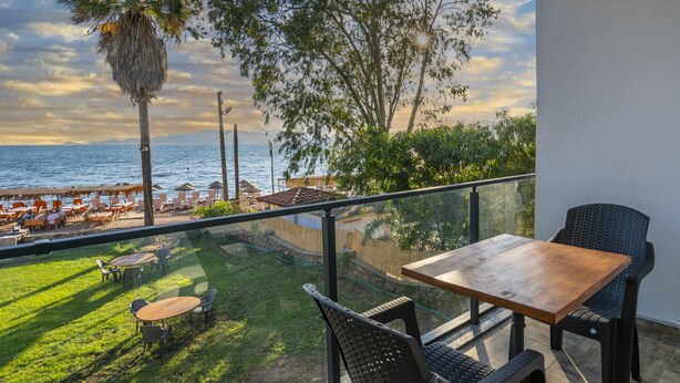 Ein Balkon mit Holztisch und Stühlen, der auf einen Strand mit Sonnenschirmen und einer Palme blickt.