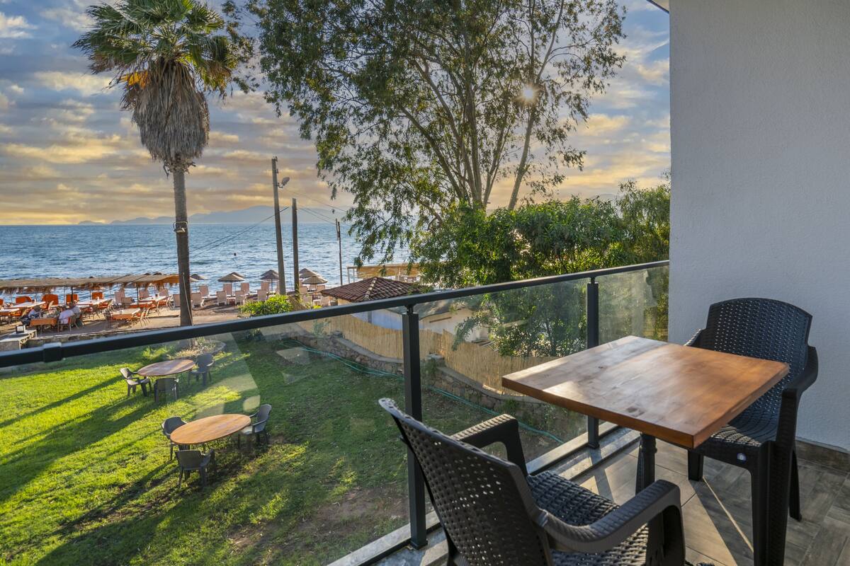 Uma varanda com mesa e cadeiras de madeira, com vista para uma praia com guarda-sóis e uma palmeira.