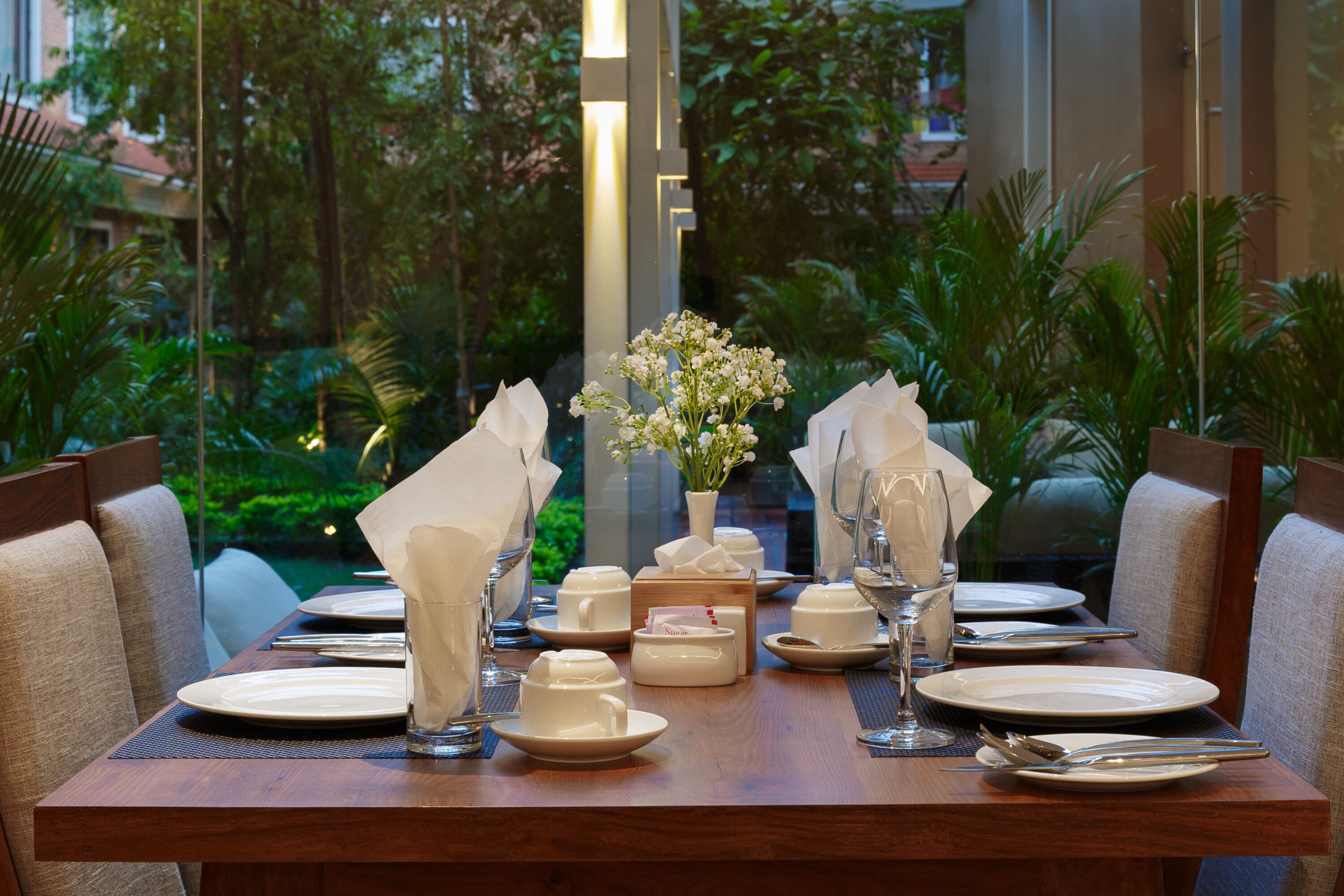 A dining table set with white plates, glasses, and napkins, surrounded by chairs, with a vase of flowers in the center.