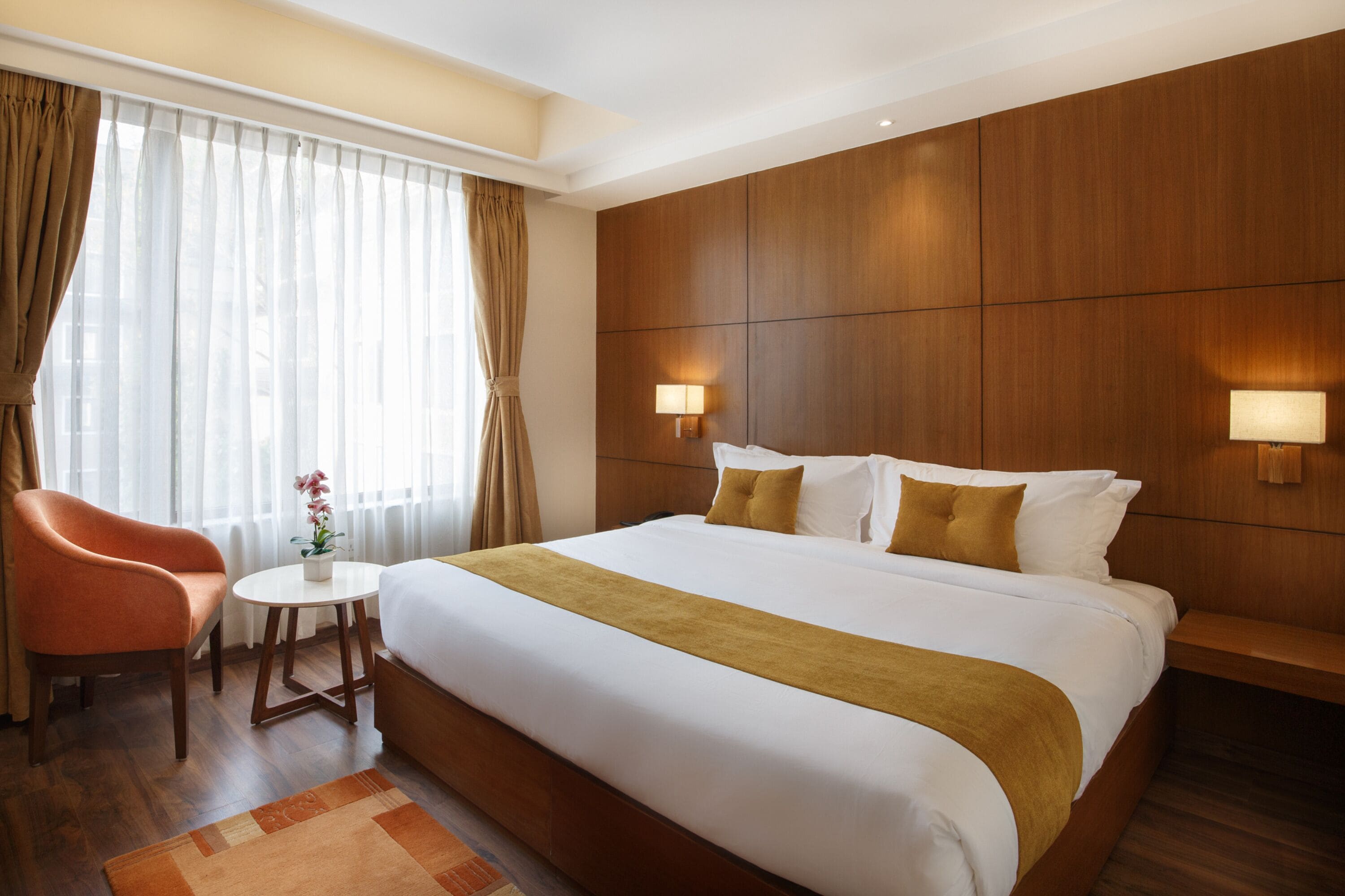 A hotel room with a large bed, wooden headboard, two bedside lamps, a small round table with a plant, and a chair.