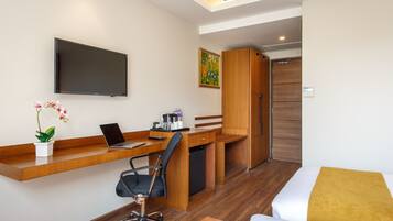A hotel room with a wooden desk, a laptop, a chair, a bed with a yellow throw, a TV, and a painting on the wall.