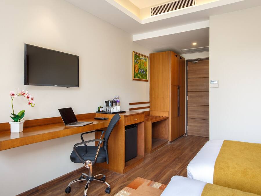 A hotel room with a wooden desk, a laptop, a chair, a bed with a yellow throw, a TV, and a painting on the wall.