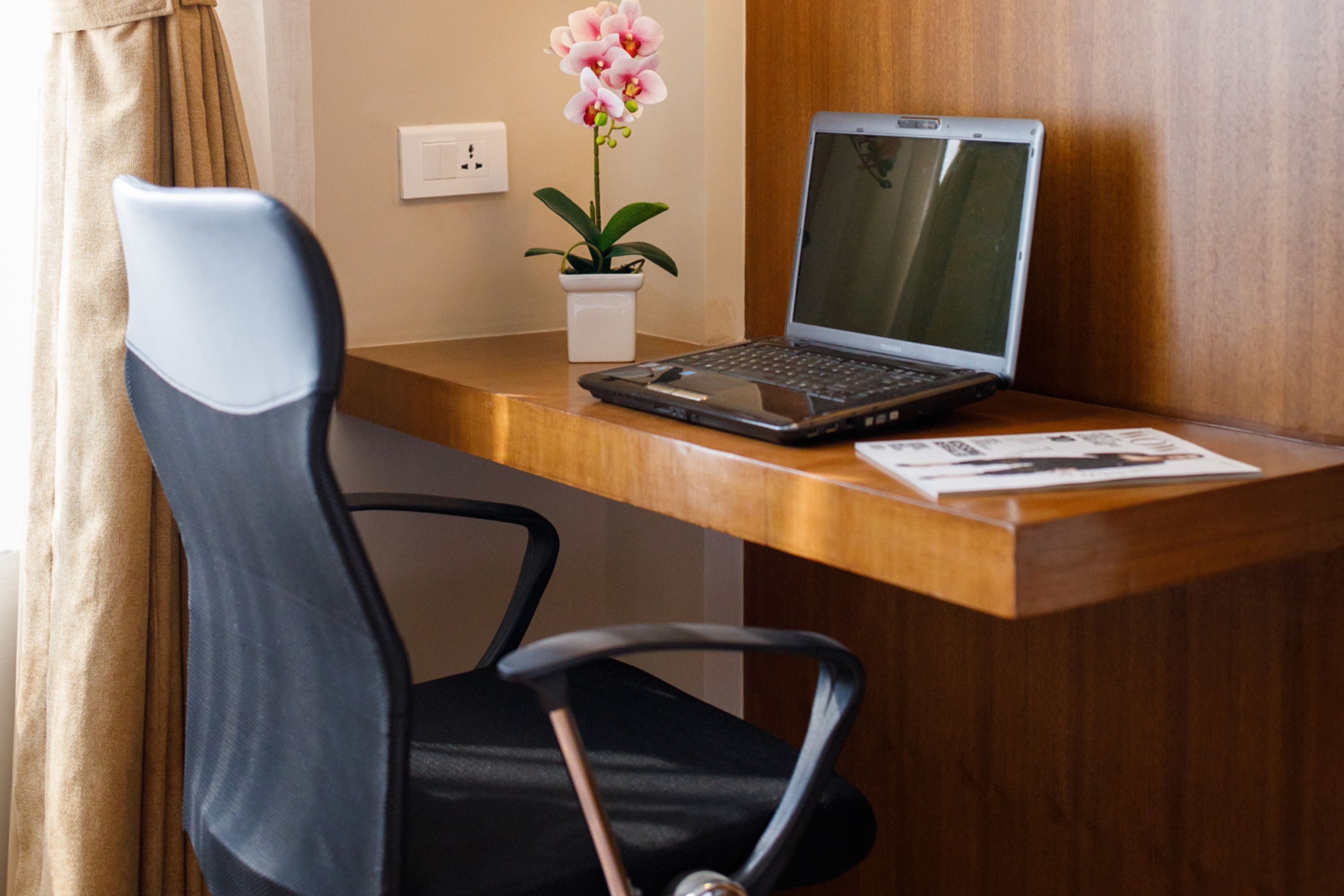 A laptop on a wooden desk with a chair, a potted orchid, and a power outlet on the wall.