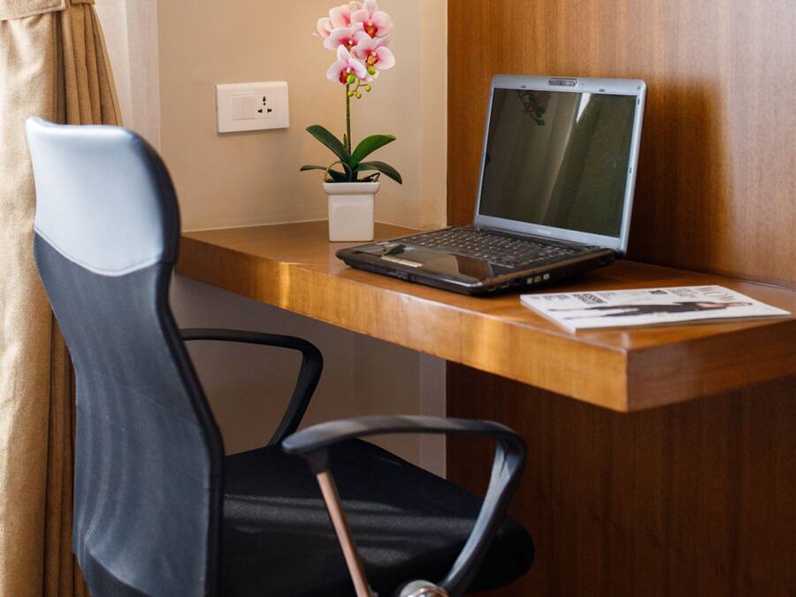 Een laptop op een houten bureau, met een stoel, een potorkidee en een stopcontact aan de muur.