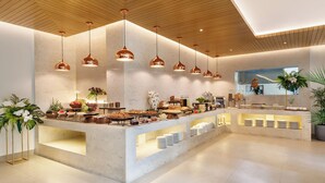 Un moderno vestíbulo de hotel con una zona de buffet central, iluminación colgante y plantas en macetas.