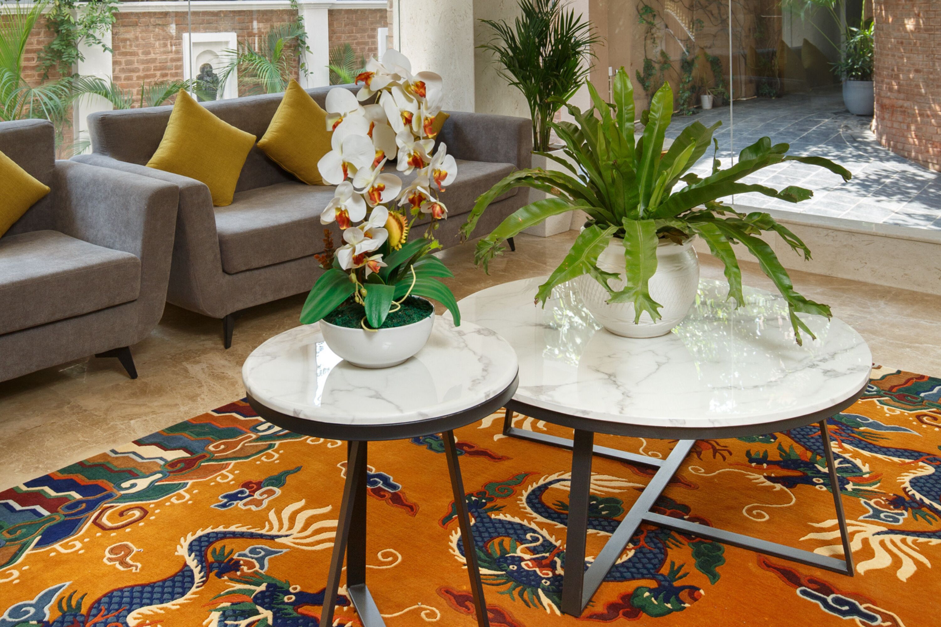 Una sala de estar moderna con un sofá gris, cojines amarillos, un tapete con estampado de dragones coloridos y dos mesas de mármol blanco con plantas en macetas.