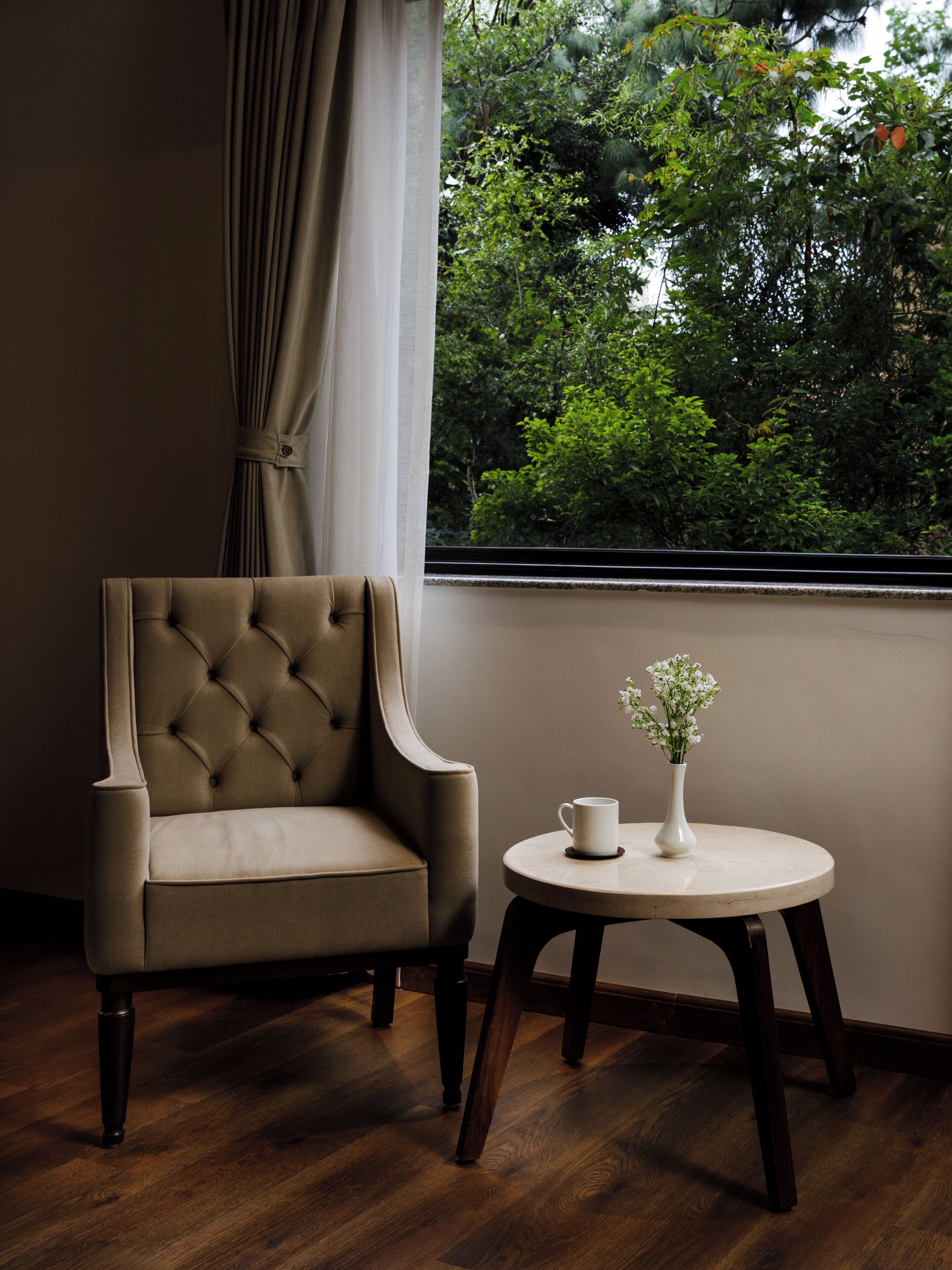 A room with a tufted armchair, a small round table with a white vase and a cup, and a window with a view of greenery.