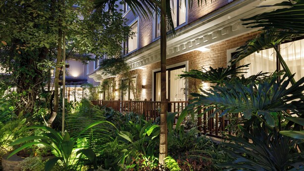 Un jardín bien iluminado con diversas plantas tropicales y un edificio de ladrillo con balcón.