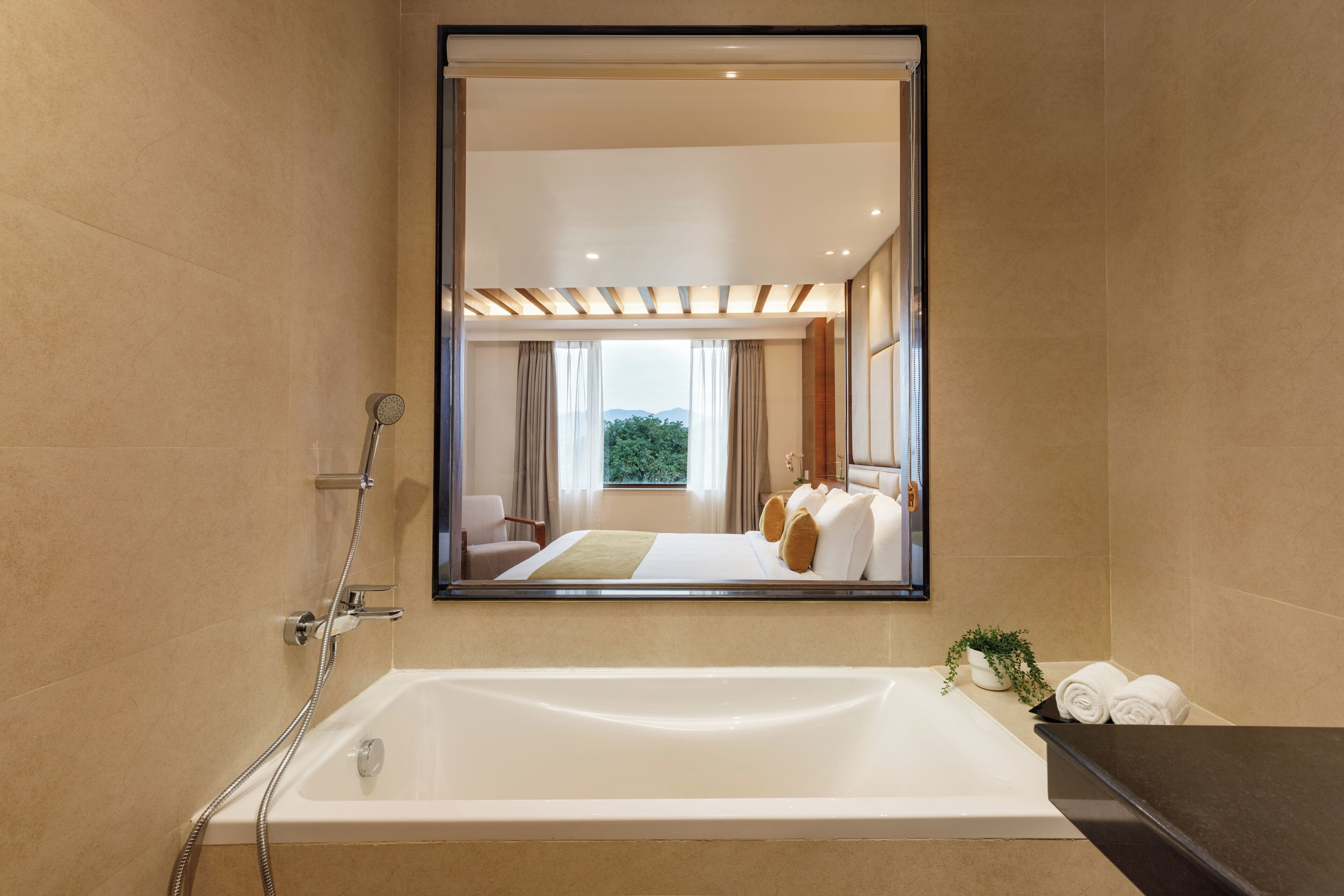 A modern bathroom with a large bathtub, a shower, and a mirror reflecting a bedroom with a bed and curtains.