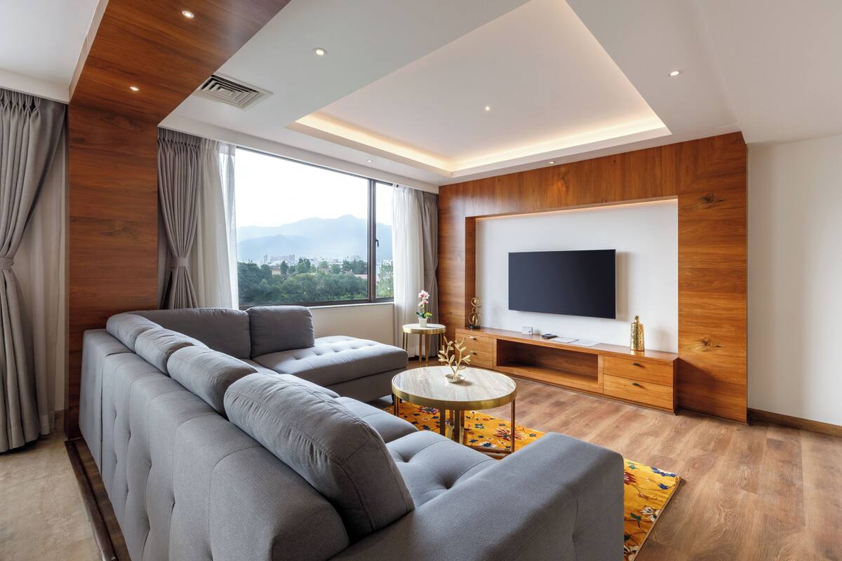 A modern living room with a sectional sofa, a round coffee table, a flat-screen TV, and a large window offering a city view.