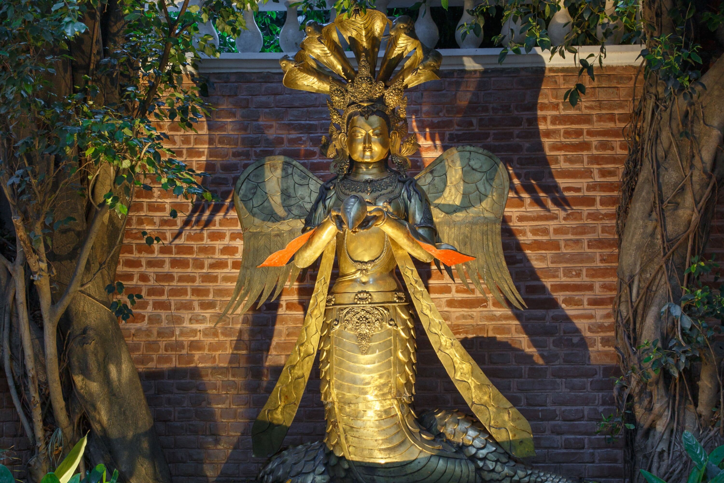 A statue of a figure with wings and a headdress, set against a brick wall with vines.