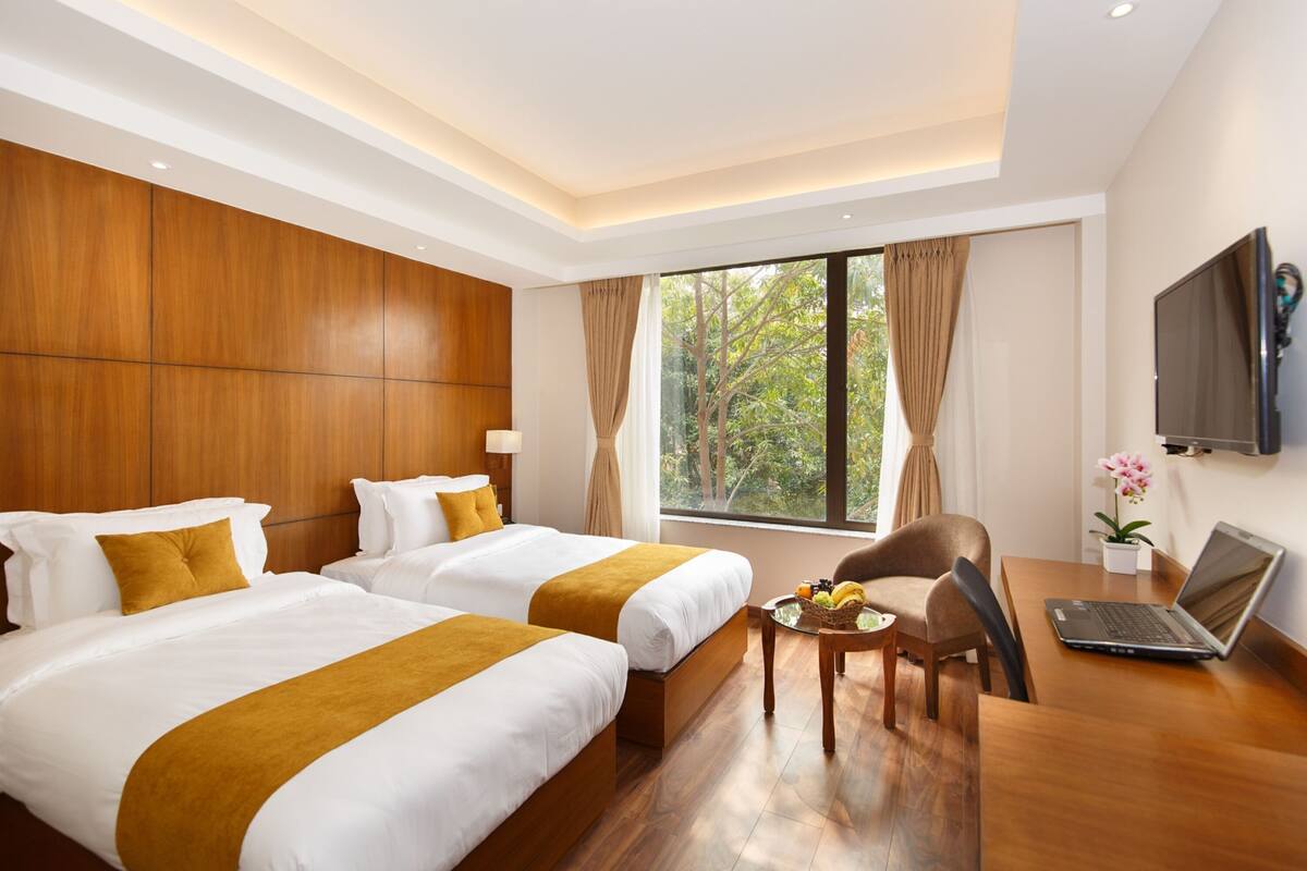 A hotel room with two beds, a desk with a laptop, a TV, and a window with curtains.