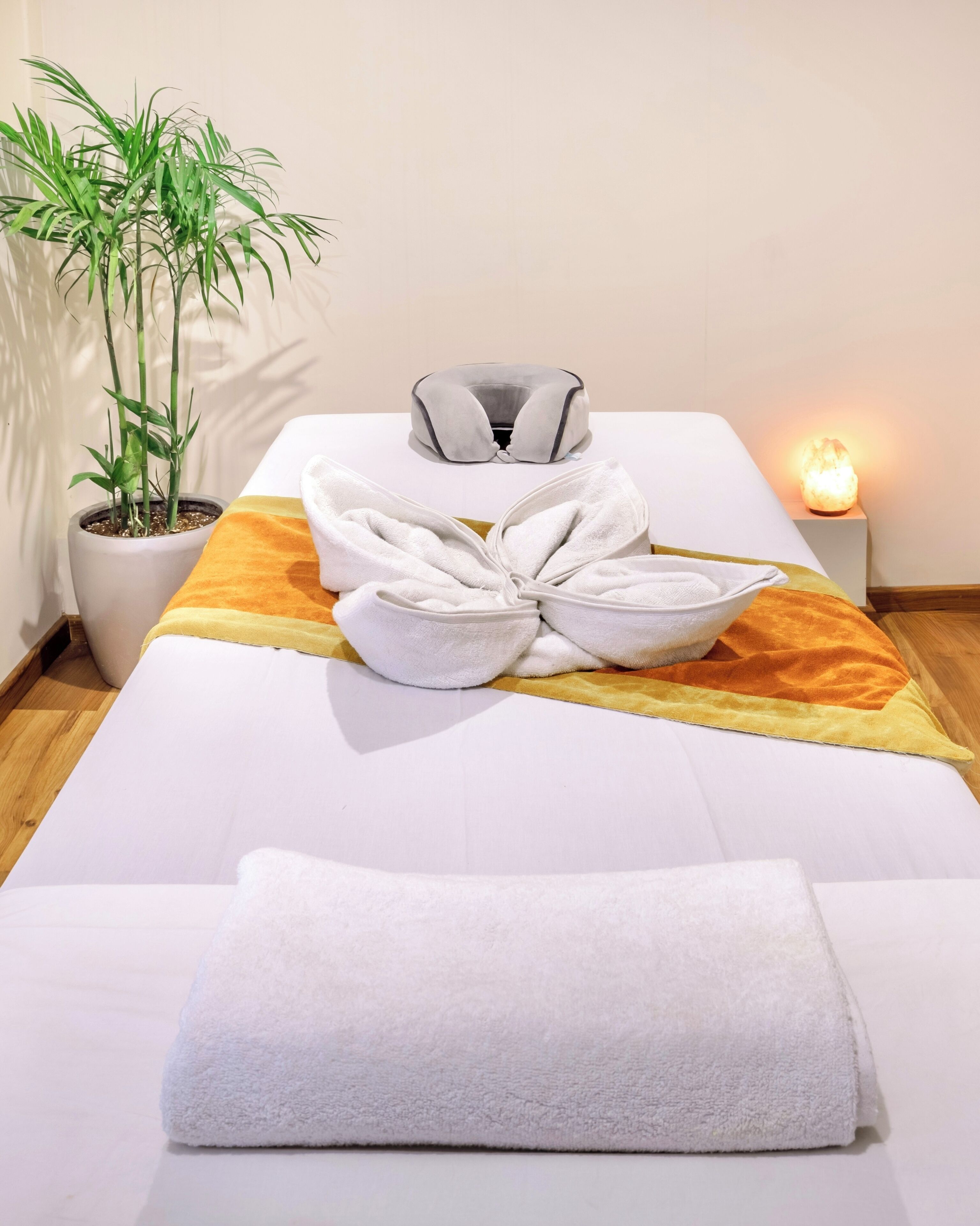 Una cama bien hecha con una toalla blanca, un arreglo decorativo de toallas, una silla de masajes, una planta en maceta y una vela encendida.