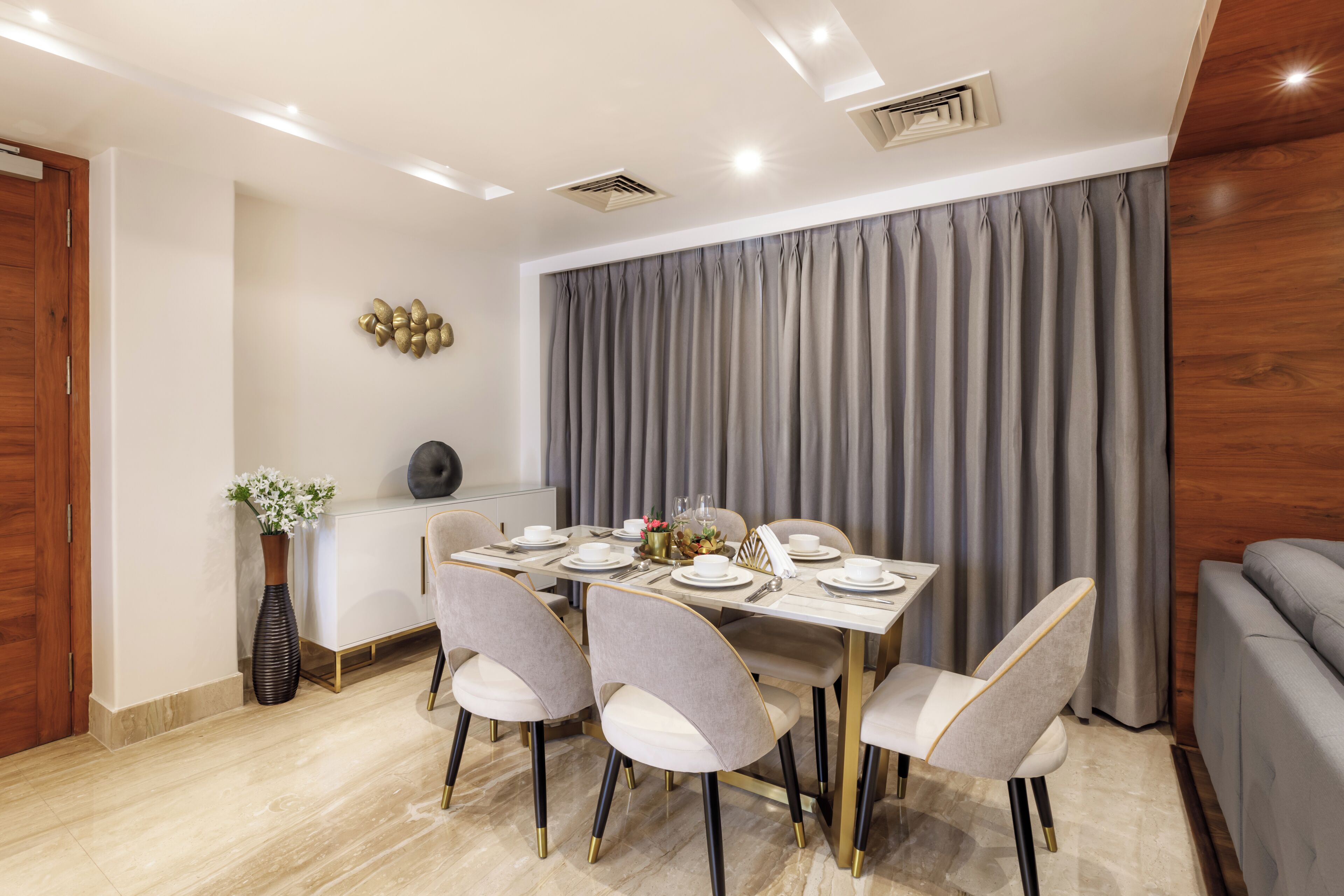A dining area with a table set for four, chairs with gold legs, a vase with flowers, and a grey curtain backdrop.