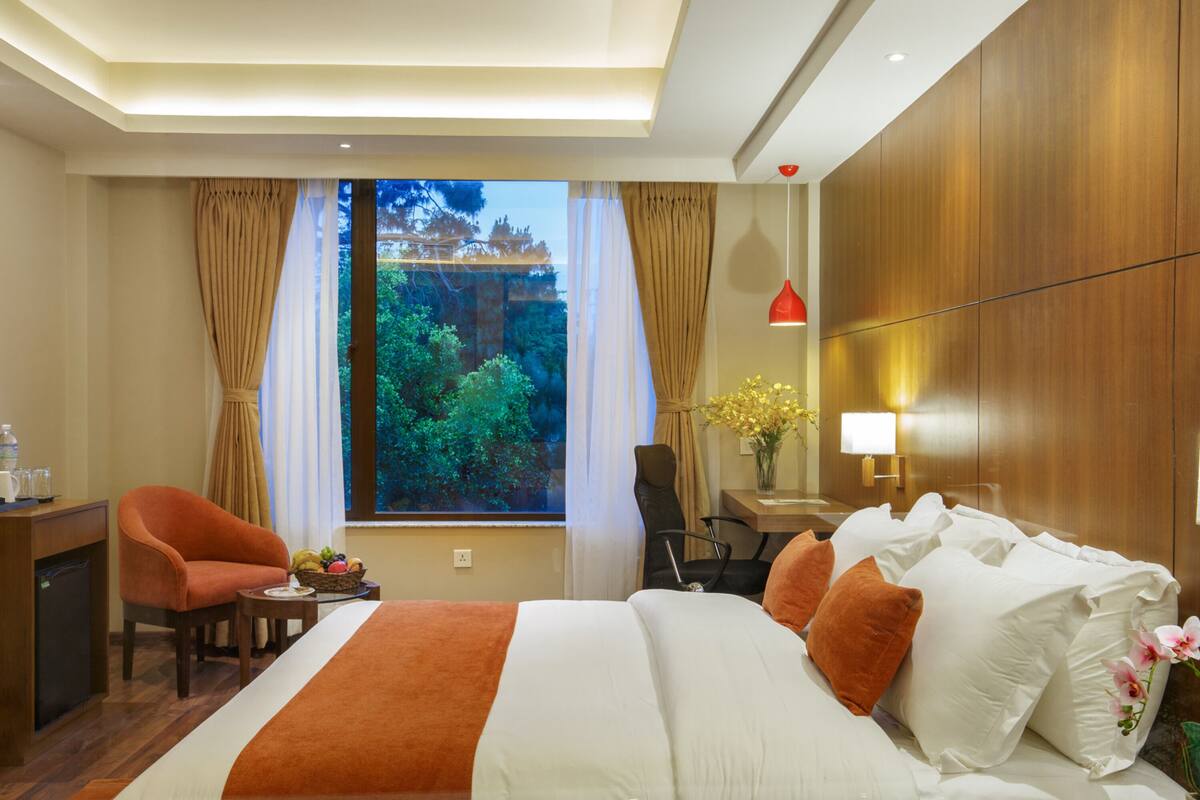 A hotel room with a large bed, a desk, and a chair. There is a window with curtains, a lamp, and a vase with flowers.